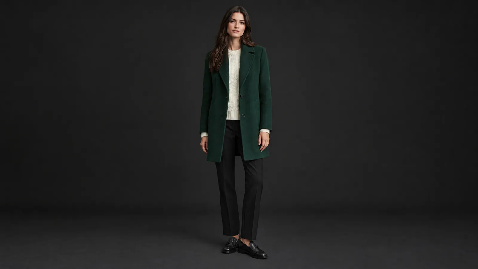 Ganzkörper-Katalogfoto eines Models vor dunkelgrauem Hintergrund in dunkelgrünem Wollmantel, cremefarbenem Strickpullover und schwarzer Hose