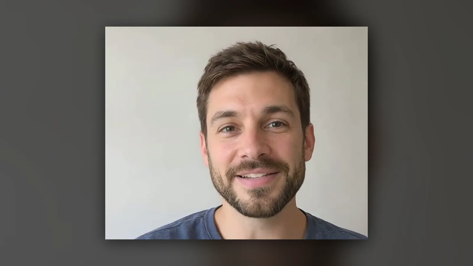 A bearded creator smiling and talking to camera in front of a light grey wall