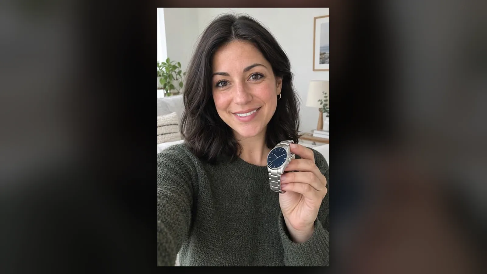 A smiling dark-haired woman taking a selfie at home while holding up a silver wristwatch